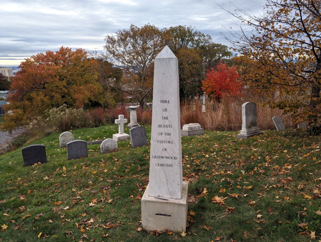 Sophie Calle installation of "Here Lie the Secrets of the Visitors of Green-Wood Cemetery," 2017. A obelisk with a box with a slot for paper beneath it.