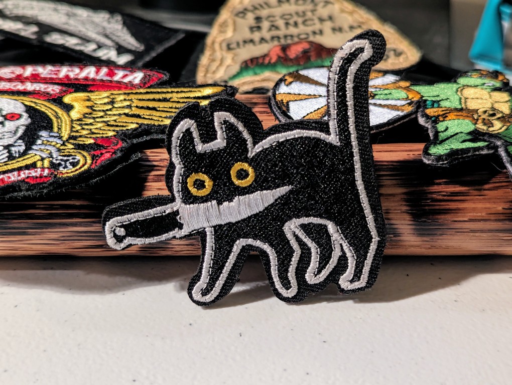 An embroidered patch of a black cat carrying a knife in its mouth. Other embroided patches are seen in the background.