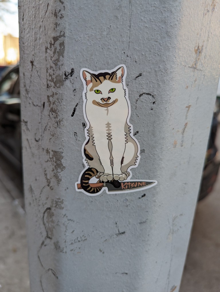 Cat standing over a knife with the words "Kitsune" on the blade. A sticker on a lightpole in Park Slope, Brooklyn.
