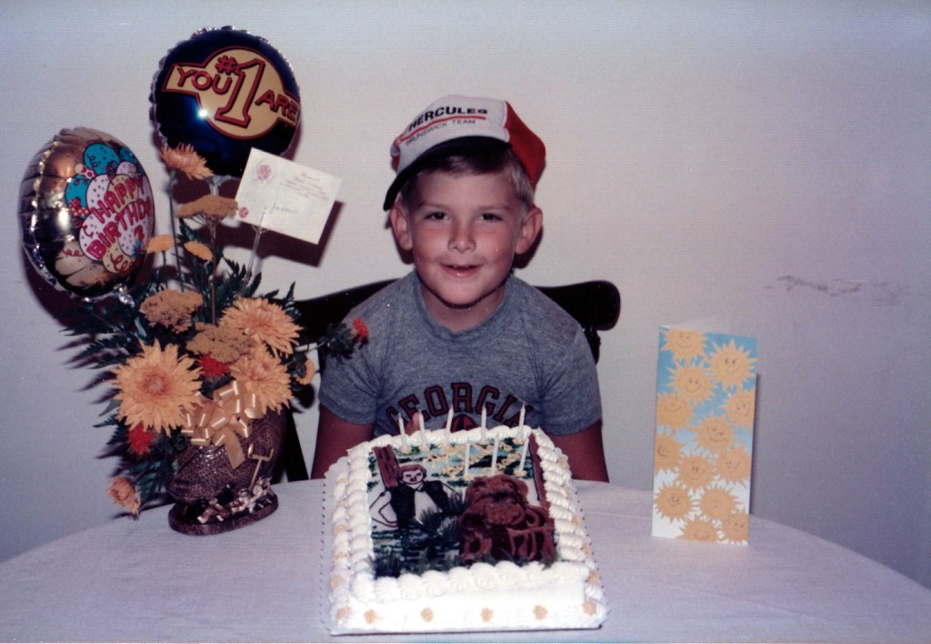 Young boy wearing a Hercules baseball cap and Georgia Bulldogs shirt sitting behind a Star Wars themed Han Solo and Chewbacca birthday cake.