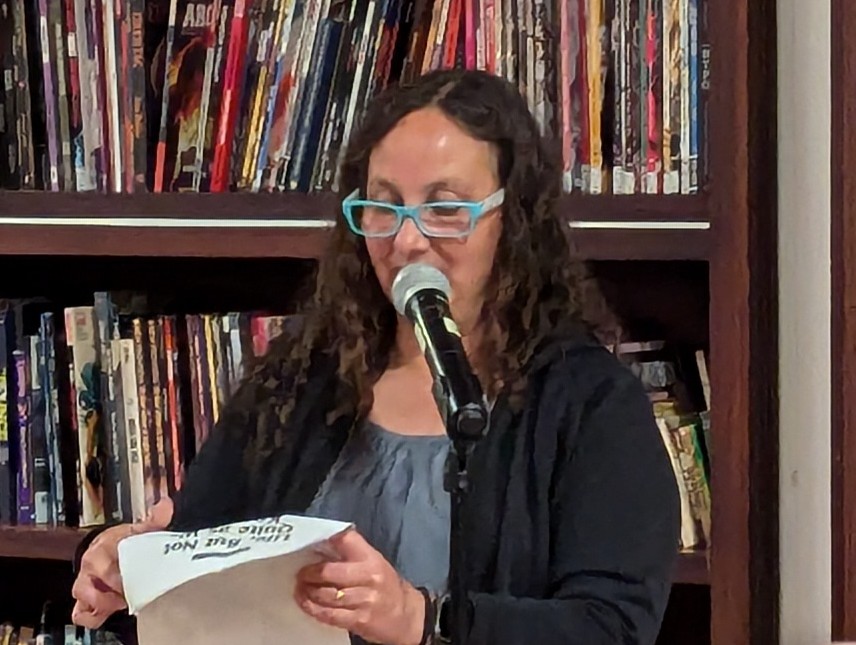 Christina De La Rocha standing by a microphone and reading from a text.