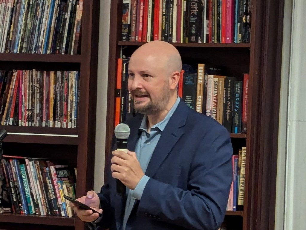 Trevor Quachri holding a microphone and reading from a text.