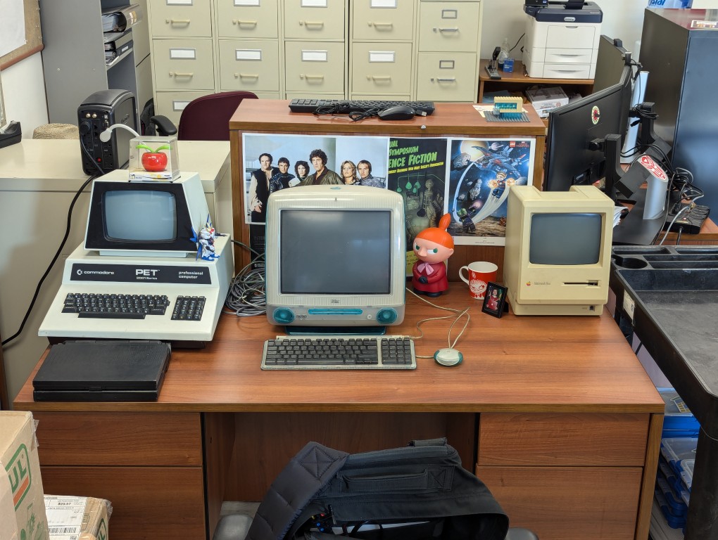 Powerbook, Commodore PET, Apple iMac, and Macintosh 512K on an office desk