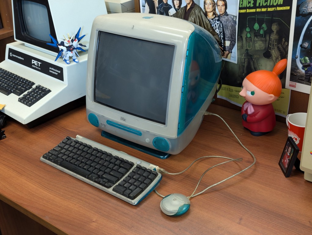 original bondi blue imac on a wood desk, whatnots and other computers on desk in background