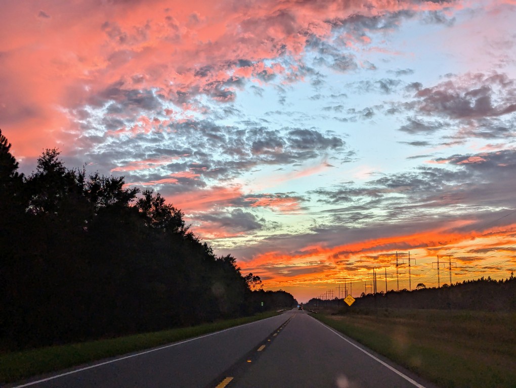 a paved two lane road extending to the horizon at dusk, the sun illuminates the clouds above in a variety of colors