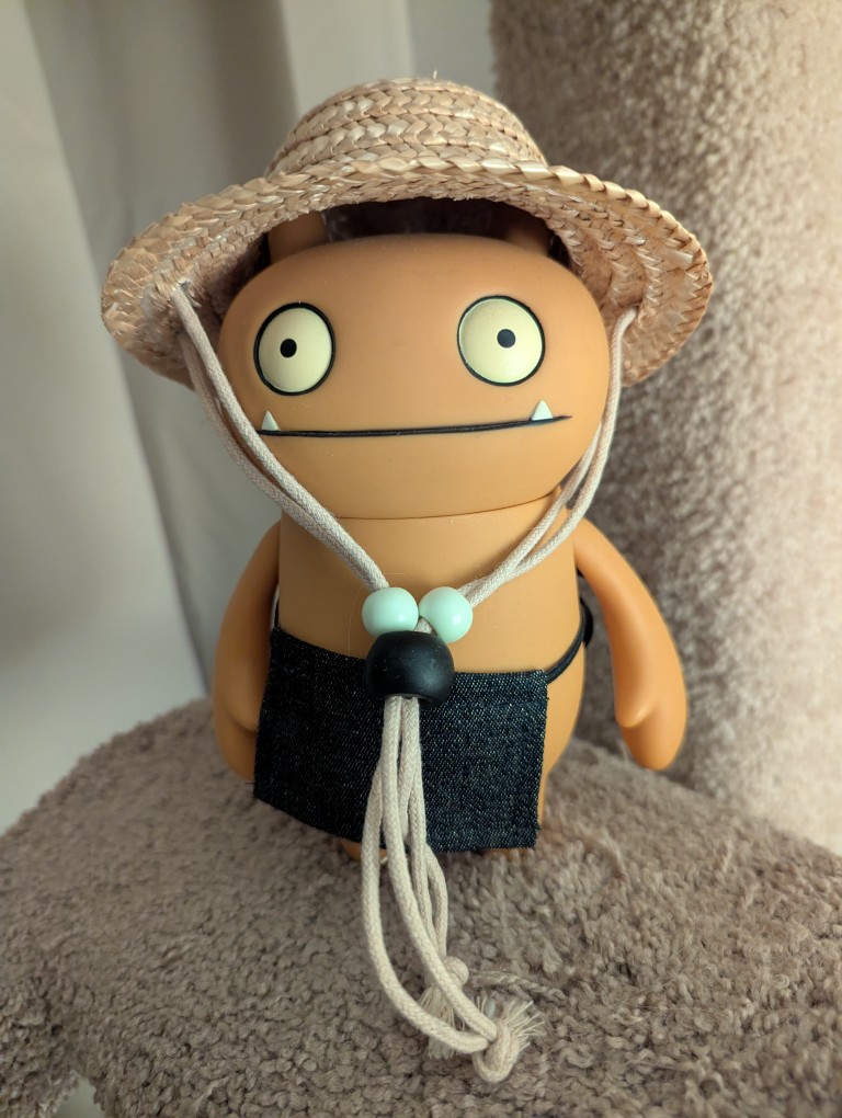 wage the uglydoll is an orange anthromorphic monster with orange skin and two tiny upturned teeth wearing a worker's apron and a straw hat with lanyard.