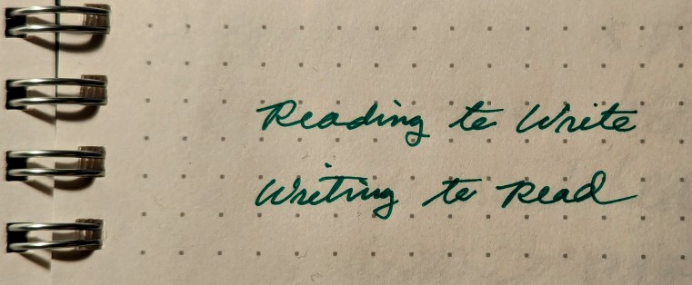 the words "Reading to Write" and "Writing to Read" written in cursive in a notebook with a dot-grid background and spiral bound