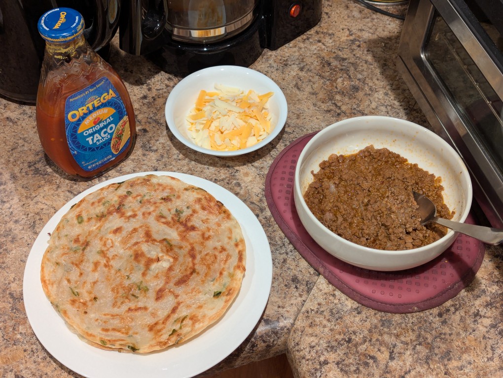 from left to right: a bottle of ortega medium taco sauce, a small bowl of shredded cheese, a large bowl of ground beef and onion taco mix, and a green onion pancake on a plate
