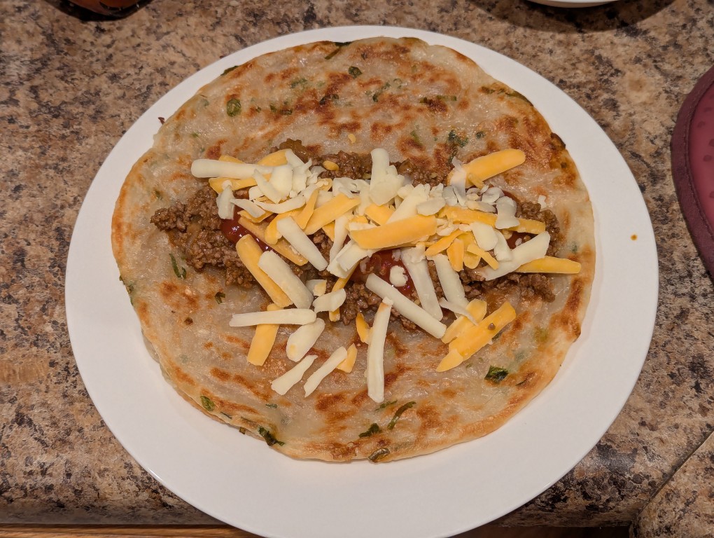 a green onion pancake flat with ground beef taco mix, sauce, and shredded cheese over a flat green onion pancake in a white ceramic plate