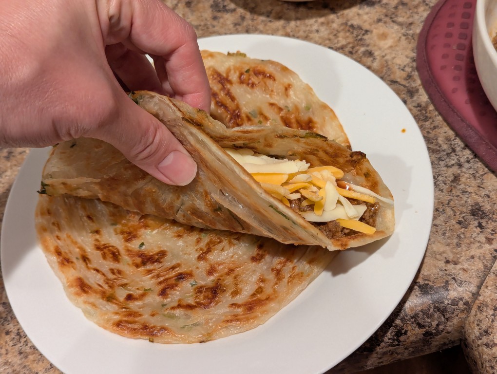 hand holding a green onion pancake folded in half filled with ground beef taco mix, sauce, and shredded cheese over a flat green onion pancake in a white ceramic plate