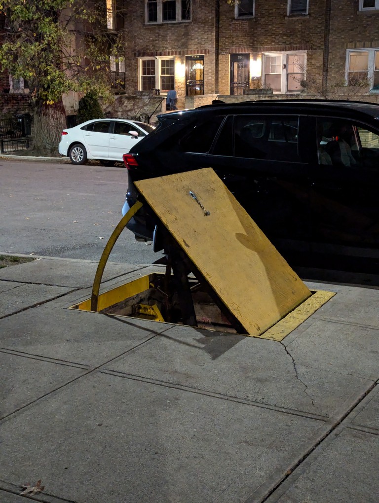 closeup of a metal cover is held open at about 45 degrees allowing access to a passageway built into the sidewalk going underground