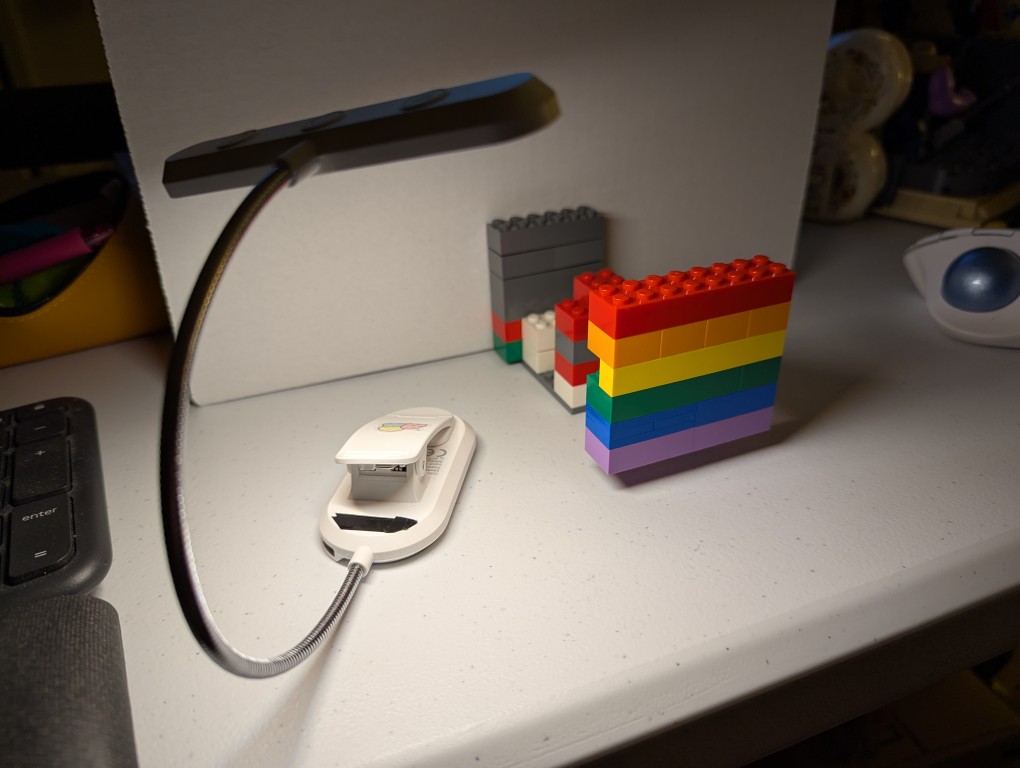lego bricks resembling the pride flag are connected to a mix of other hidden lego bricks in front of a white cardboard box and illuminated by a book reading light