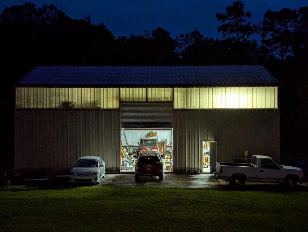 a metal building workshop at night illuminated from within, 2 cars, a truck, and jeep are seen around or inside the building