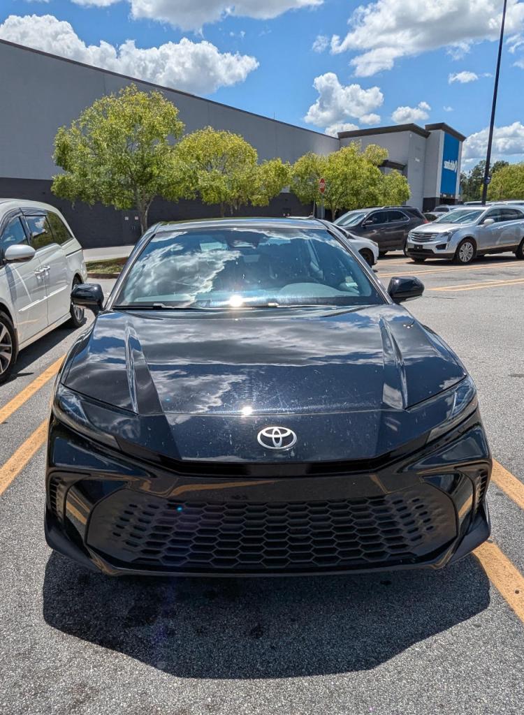 3/4 view of black late model toyota camry in a parking spot surrounded by other cars and shopping carts in background, trees with green leaves and building in the background, blue sky with clouds above