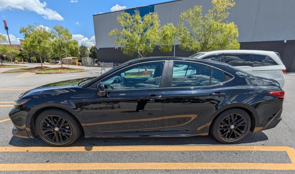side view of black late model toyota camry in a parking spot surrounded by other cars and shopping carts in background, parking lot ringed with trees with green leaves, building in background, blue sky with clouds above