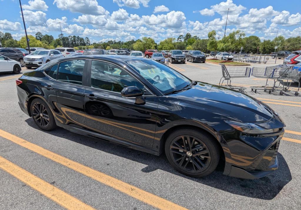 3/4 view of black late model toyota camry in a parking spot surrounded by other cars and shopping carts in background, parking lot ringed with trees with green leaves, blue sky with clouds above