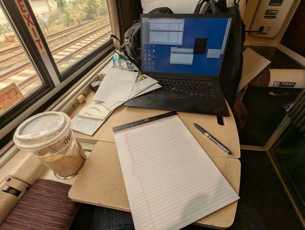 table folded out holding a writing tablet, pen, and laptop computer, train tracks are seen outside the window