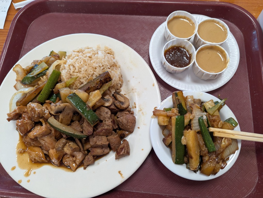 several plates: one plate of plain fried rice, hibachi steak and mushrooms, teriyaki chicken, stirfried zucchini, a bowl with extra stir fried veggies, and a small plate with three cups of mustard sauce and one cup of ginger sauce