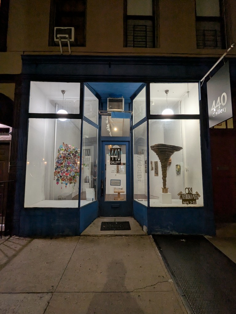 high glass entrance to 440 gallery. a colorful assemblage artwork on the right and a cardboard tornado artwork on the right. it's night but the lights are on inside