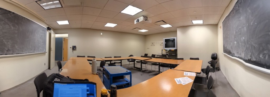 panoramic view of a seminar style classroom from a corner opposite the door, two chalkboards, overhead projector, tv in back