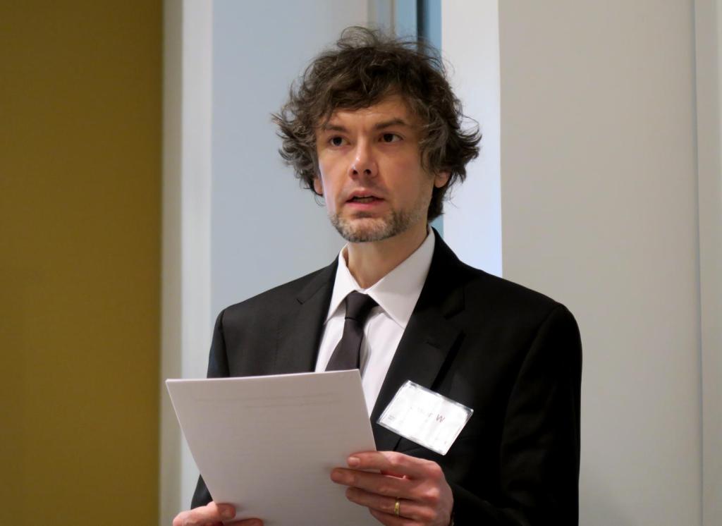 while male with messy dark hair in black suit speaking at a conference holding papers