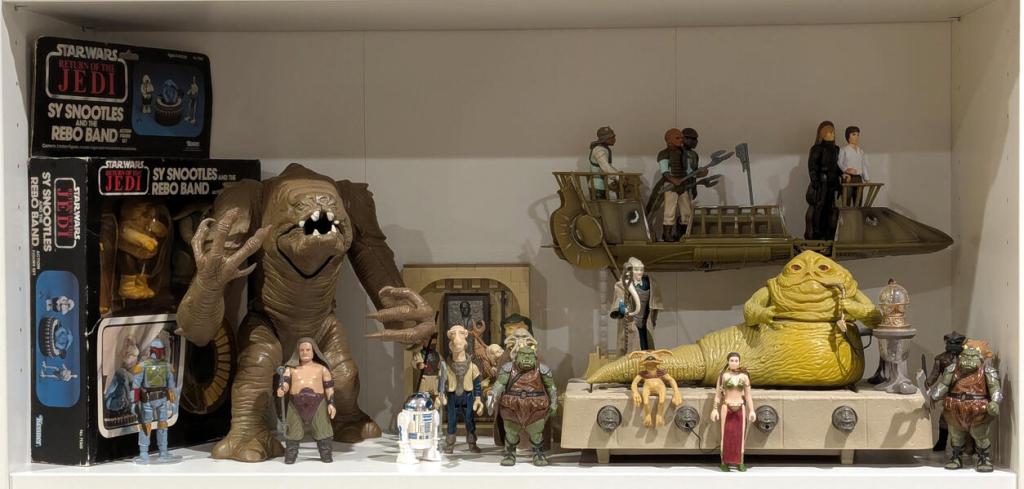 Star Wars vintage and modern action figures and vehicles on a bookshelf and desk.